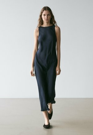 Woman walking forward wearing a sleeveless navy blue midi dress and black flats against a plain light gray background.