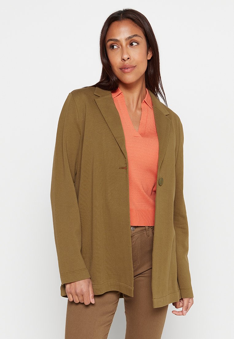 Olivgrüner Blazer mit einem einzigen Knopf, taillierte Passform und mit Schößchen, kombiniert mit einem orangefarbenen V-Ausschnitt-Oberteil und braunen Hosen.
