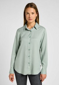 Chemise à manches longues en tissu vert clair, dotée d'un col classique, d'une fermeture à boutons sur le devant et d'un ourlet arrondi. Texture lisse et poignets standards.