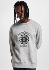 Uomo con dreadlocks che indossa un felpa grigia con la scritta "University League" e un logo Jack & Jones, in piedi contro uno sfondo bianco uniforme.