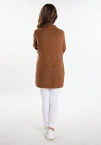 Gezellige bruine lange trui van chunky breiwerk, met een hoge kraag en een licht oversized pasvorm, gecombineerd met witte jeans en witte schoenen.