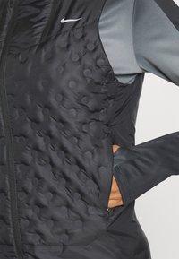 Chaleco sin mangas negro de Nike con textura acolchada y patrones circulares, acentos de tela gris y un bolsillo con cremallera en el lateral.