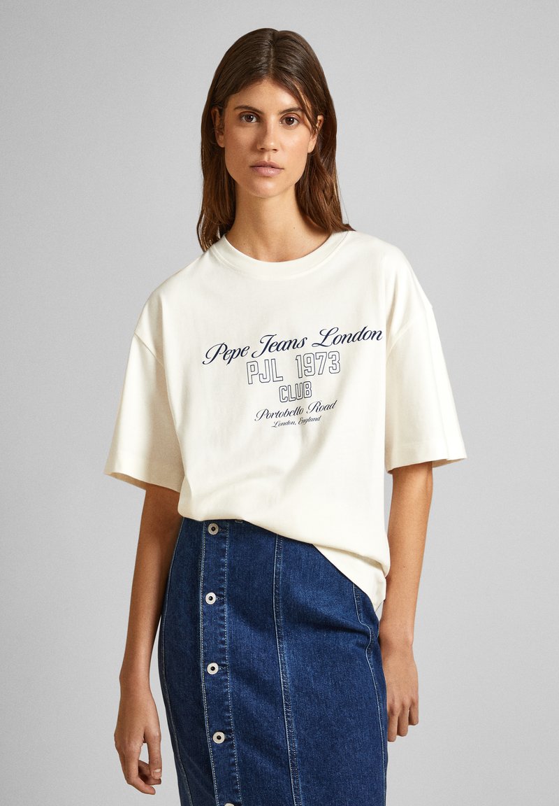 Cremige Baumwoll-T-Shirts mit kurzen Ärmeln, auf denen der Text "Pepe Jeans London" in Marineblau und Weiß zu sehen ist. Kombiniert mit einem blauen Jeansrock, bei dem die Knopfdetails sichtbar sind.