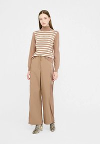 Braun gestreifter Pullover mit hohem Kragen, kombiniert mit beige hoch taillierter, weit geschnittener Hose und gemusterten Stiefeletten mit Absatz.