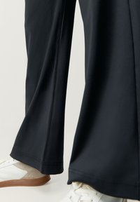 Čierne široké nohavice z hladkého materiálu, so šitými švami, v kombinácii s bielymi teniskami a béžovým akcentom.