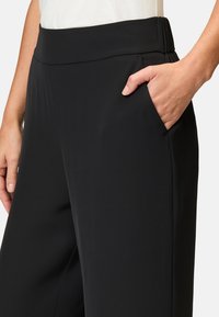 Pantalons noirs en tissu lisse et extensible, avec une large taille élastique et des poches latérales. Le design est ajusté et épuré.