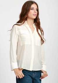 Camicia bianca a maniche lunghe con bottoni, dotata di due tasche sul petto, realizzata in un tessuto leggero e trasparente. Presenta un colletto classico e maniche con polsini.