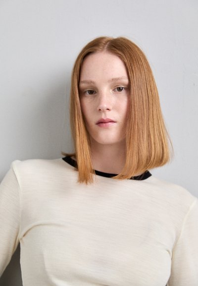 Korte mouwen, crèmekleurig shirt met een zwarte kraag, met een gladde textuur en een licht losse pasvorm. Model heeft steil, middel-lang oranje haar.