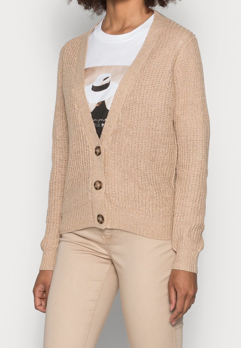 Personne portant un cardigan en maille beige avec trois boutons sur un t-shirt blanc à motif graphique et un pantalon beige, sur un fond uni.