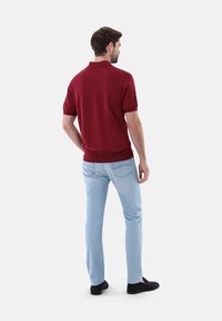 Burgunderfarbenes Polohemd mit kurzen Ärmeln, tailliertem Schnitt und geripptem Saum, kombiniert mit hellblauen Jeans und schwarzen Schuhen. Die Rückansicht zeigt einen schlichten Stil.