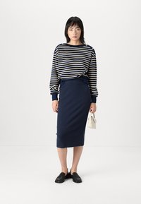 Pull en laine rayé bleu marine et crème à manches longues, jupe bleu marine taille haute, chaussures noires à enfiler, tenant un sac à main blanc. Design simple et ajusté.