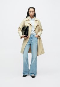 Béžový trenčkot na gombíkovanej prúžkovanej košeli, v kombinácii so svetlomodrými zvonovými džínsami a čiernymi lodičkami so zašpicatenou špičkou. V ruke čierna listová kabelka.