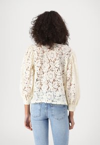 Blusa in pizzo crema con maniche a palloncino, caratterizzata da motivi floreali. La parte posteriore è parzialmente trasparente e il fondo è aderente.