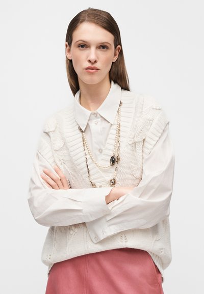 Gilet blanc en maille avec motifs texturés porté sur une chemise blanche à col. Manches longues avec poignets à boutons. Jupe rose et colliers superposés.