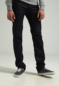 Jeans in denim scuro con una vestibilità dritta, dotati di un girovita medio e abbinati a sneaker grigie. Texture liscia e dettagli di cucitura sottili.