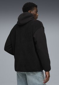 Schwarze Fleecejacke mit einer strukturierten, plüschigen Rückseite und glatten Ärmeln, ausgestattet mit einer Kapuze. Kombiniert mit hellblauen Jeans.