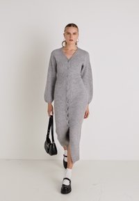 Abito cardigan grigio con maniche lunghe, scollo a V, frontale con bottoni, lunghezza a metà polpaccio, texture a coste, abbinato a una piccola borsa nera e scarpe.