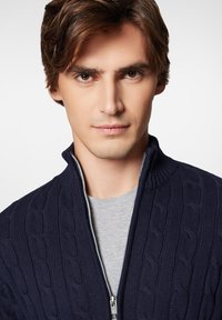 Granatowy sweter z dzianiny warkoczowej z zamkiem błyskawicznym i wysokim kołnierzem, noszony na jasnoszarym T-shircie; ma wykończenia ze ściągacza oraz srebrny zamek.