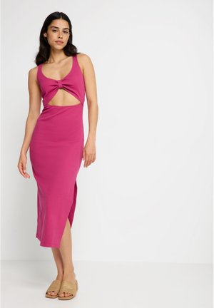 Woman wearing a sleeveless magenta dress with a front cutout and knot detail, paired with beige woven slide sandals, standing against white background.