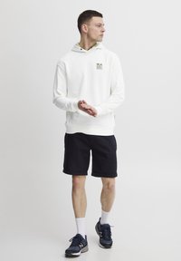 Hvit hettegenser med logo foran, sammen med svarte shorts. Modellen har på seg marineblå joggesko og hvite sokker, stående i en neutral stilling.