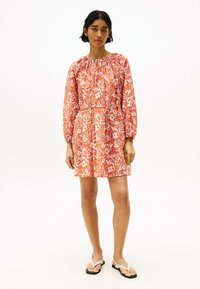 Robe à motif floral en orange et blanc, avec manches longues, encolure froncée et coupe décontractée. Portée avec des tongs.