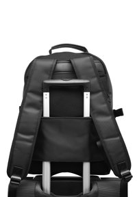 Mochila negra con correas acolchadas, detalles de malla y un panel trasero diseñado para acomodar las asas del equipaje. Textura suave, cierre de cremallera.