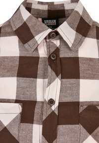 Camicia a quadri marrone e bianca realizzata in tessuto intrecciato, caratterizzata da un colletto a punta, due bottoni e una tasca sul petto sul lato sinistro.