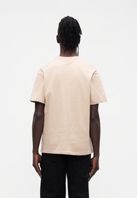 Beżowy bawełniany t-shirt z krótkim rękawem, okrągłym dekoltem i luźnym krojem, noszony z czarnymi spodniami. Bez widocznych wzorów ani metalowych detali.