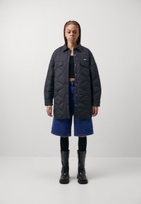 Black quilted jacket with a button front and two chest pockets, worn over a black crop top, paired with blue denim shorts and black boots.