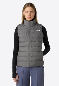 Nainen, jolla on yllään harmaa toppausliivi ilman hihoja, jossa on korkea kaulus, musta pitkähiha, ja siniset housut, seisoo yksi käsi taskussa.