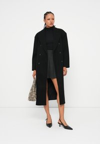 Manteau long noir à double boutonnage avec un col cranté, porté sur un haut noir et une jupe grise, associé à un sac à motif serpent et des talons noirs.