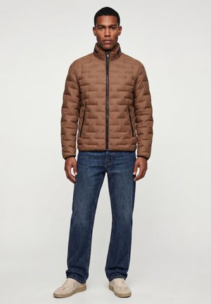 Mann steht, trägt eine braune gesteppte Jacke, blaue Jeans und hellbeige Sneakers vor einem schlichten weißen Hintergrund.