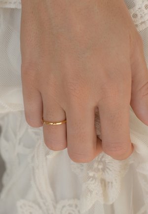 Goldring an einer Hand mit strukturiertem Finish, bestehend aus einem schmalen Band und einer glatten, polierten Oberfläche. Die Hand liegt auf einem Stoff mit Spitzenverzierungen.