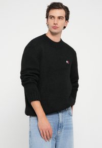 Czarny sweter wykonany na drutach z okrągłym dekoltem, ribbowymi mankietami i małą naszywką z logo w kolorach czerwonym, białym i niebieskim na lewej piersi.