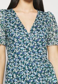 Květinové šaty s obalem v modré a zelené barvě. Mají krátké balónové rukávy, hluboký V-výstřih a lehký, průhledný materiál. Jsou zdobeny malými květinami.