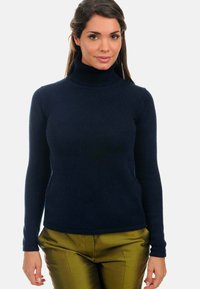Pull à col roulé bleu marine avec texture côtelée et manches longues, associé à un pantalon vert olive brillant, sur fond neutre.