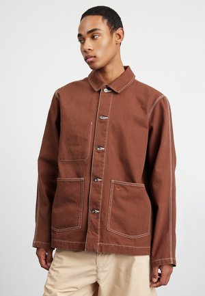 Veste en toile marron avec un col, boutons sur le devant, deux grandes poches, des coutures blanches contrastantes et un petit logo.