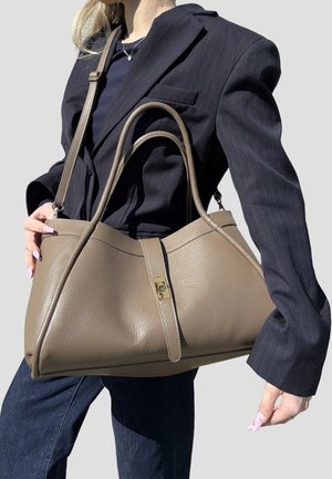 Donna con blazer nero a righe sottili e jeans scuri, che tiene in mano una grande borsa in pelle taupe con doppi manici e chiusura dorata.