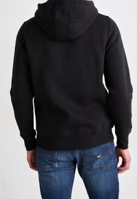 Zwarte hoodie met een trekkoordkap, geribbelde manchetten en tailleband, gecombineerd met blauwe jeans met een patchzakdetail. Zacht, katoenen materiaal.