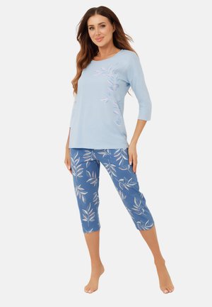 Kobieta ubrana w jasnoniebieską bluzkę z rękawami do połowy przedramienia z motywem liści oraz dopasowane niebieskie spodnie o długości 7/8 z wzorem liści, stojąca boso.