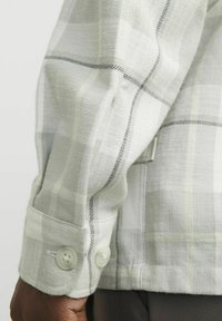 Manga de camisa de franela a cuadros en gris y blanco con acentos en verde claro. Presenta dos botones blancos en el puño y un tejido texturizado.