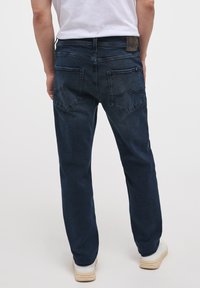 Jeans de mezclilla azul oscuro con corte recto, que presentan un ligero desgastado y bolsillos traseros clásicos, combinados con zapatillas blancas.
