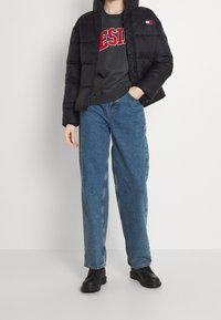 Black puffer jacket with hood, featuring a logo patch. Inside, a gray sweatshirt with red lettering. Loose-fitting blue jeans. Black chunky boots.