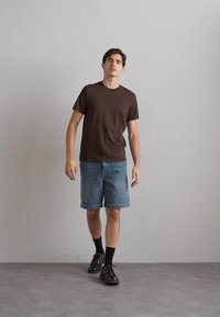 Brun kortermet t-skjorte, blå denimshorts, svarte sokker og svarte sko. Modellen står på et grått gulv mot en enkel vegg.