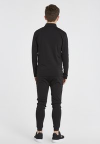 Schwarzes Sportset bestehend aus einem langärmeligen Oberteil mit Kragen und schmalen Hosen. Glatte Textur mit elastischen Bündchen und klassischen Sneakers.