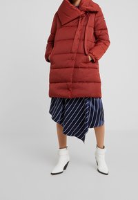Cappotto puffer rosso con un ampio colletto a fiocco e texture trapuntata. Abbinato a una gonna asimmetrica a righe blu e stivali alla caviglia bianchi con tacco.