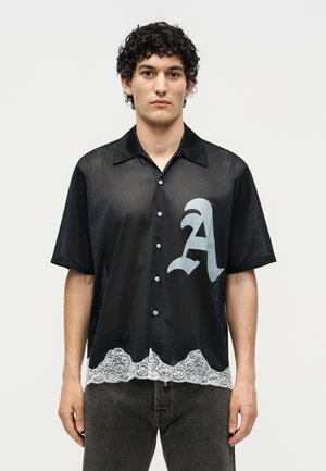 Junger Mann mit lockigem Haar, der ein schwarzes Netzhemd mit Knöpfen trägt, mit einem großen "A" und weißem Spitzenbesatz am Saum.