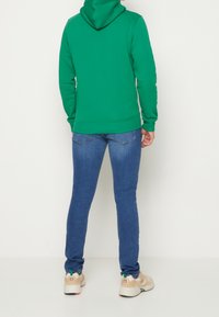 Vihreä hupullinen collegepaita, valmistettu pehmeästä kankaasta, yhdistetty istuviin sinisiin farkkuihin. Tennarit ovat beige-vihreät, kuvioiduin pohjin.