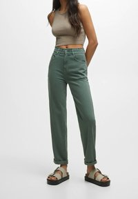 Pantalon en mélange de coton vert à coupe décontractée, taille élastique et revers roulés, associé à des sandales gris clair ornées d'un nœud décoratif.