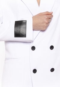 Osoba poprawiająca białą, podwójnie zapinaną marynarkę z czterema czarnymi guzikami oraz czarną prostokątną naszywką na rękawie z napisem "PILAWSKI."